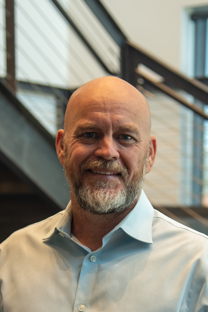 A bald man with a beard and mustache, wearing a light blue collared shirt, stands indoors in front of a staircase with metal railings—an ideal look for someone in the Precision Sheet Metal Manufacturing industry.