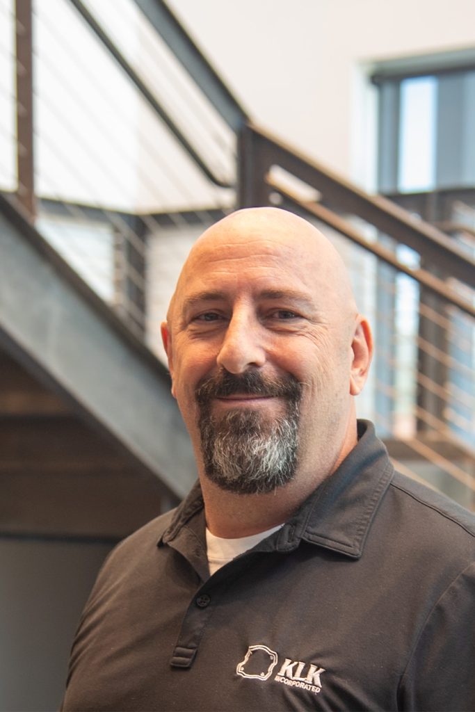 A man with a shaved head and goatee, wearing a black polo shirt with a Precision Sheet Metal Manufacturing company logo, stands indoors in front of a staircase.