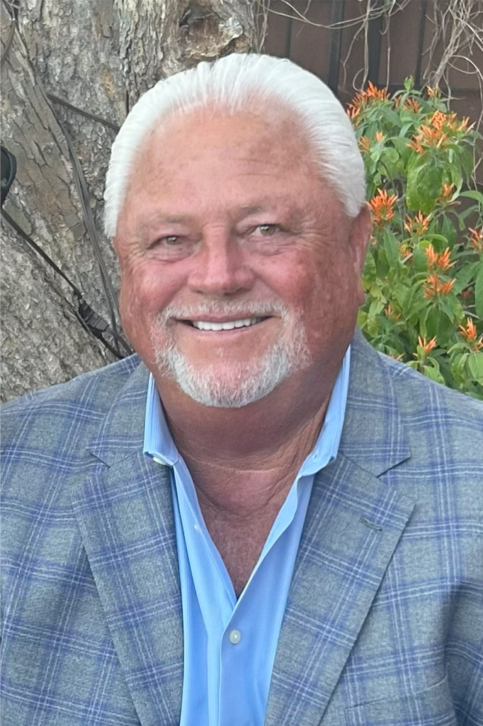 An older man with white hair and a beard, wearing a blue plaid jacket and light blue shirt, smiles while sitting outdoors near a tree and flowers, reflecting on his years in Precision Sheet Metal Manufacturing.
