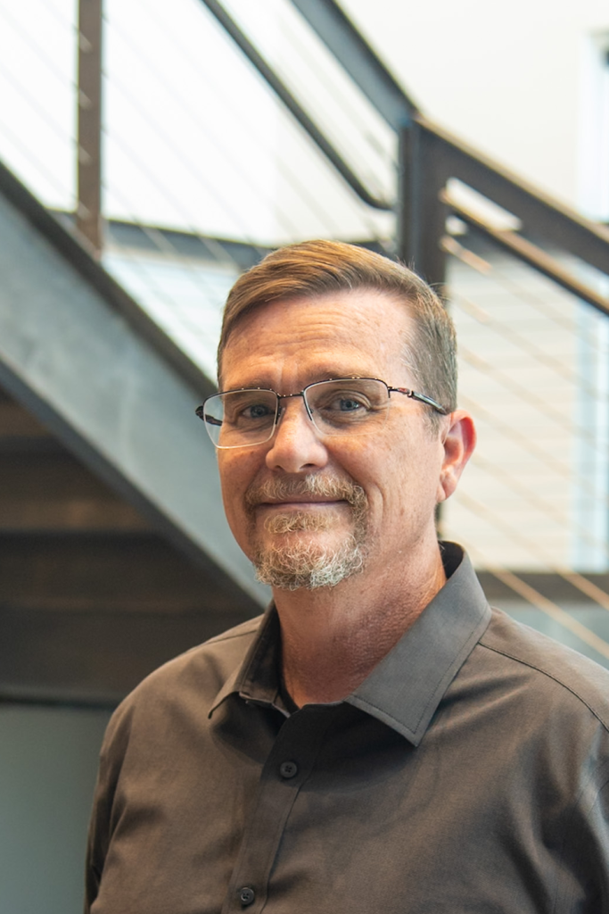 A middle-aged man with glasses, short brown hair, and a goatee stands indoors in front of a modern staircase, wearing a dark collared shirt—reflecting the professionalism of Precision Sheet Metal Manufacturing.