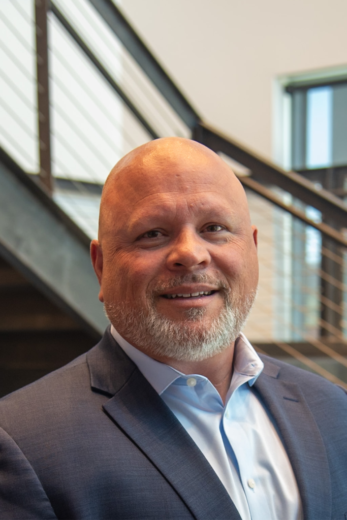 A man with a shaved head and trimmed beard, dressed in a blue suit jacket and light blue shirt, stands indoors in front of a staircase—reflecting the professionalism often found in Precision Sheet Metal Manufacturing.