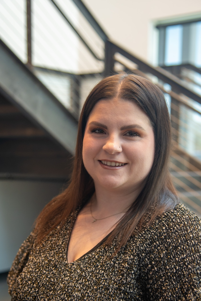 A woman with straight brown hair and a nose piercing stands indoors, smiling at the camera in front of a staircase with metal railings, echoing the modern aesthetic often seen in precision sheet metal manufacturing.