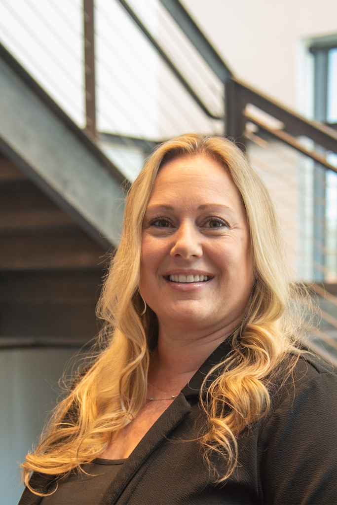 A woman with long blonde hair, wearing a black blazer, smiles at the camera indoors with a staircase in the background, embodying professionalism often seen in Precision Sheet Metal Manufacturing environments.