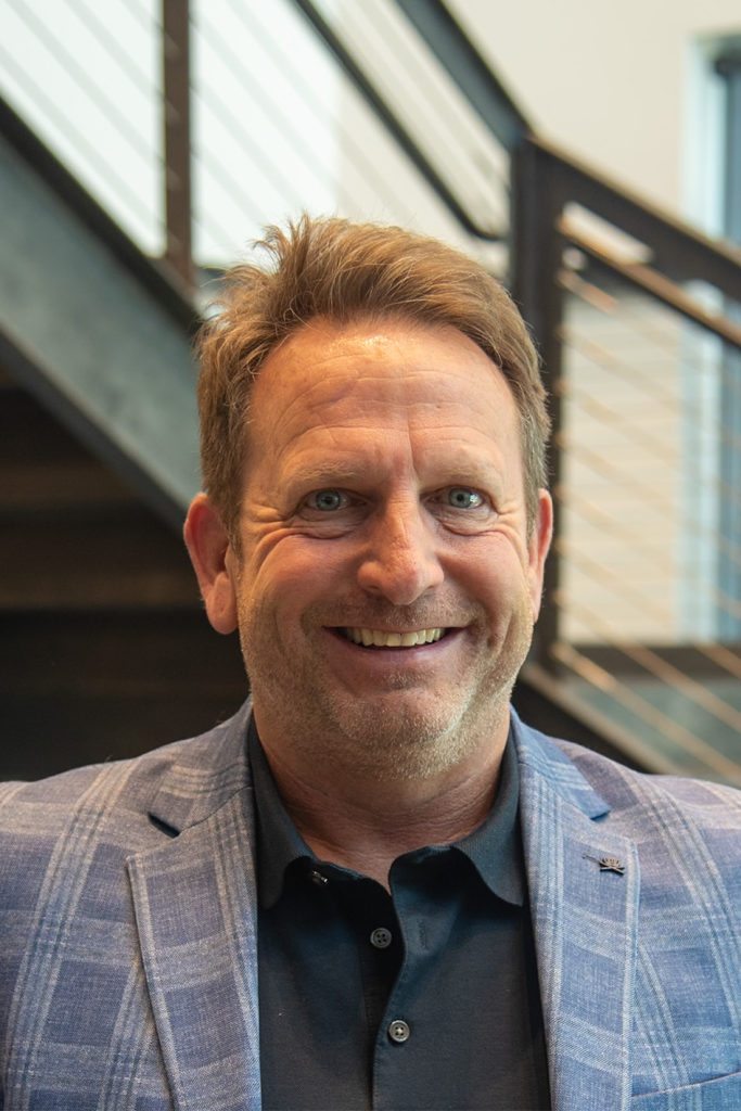 A man with light brown hair and a beard, wearing a blue plaid jacket and black polo shirt, smiles while standing indoors near a staircase at a Precision Sheet Metal Manufacturing facility.
