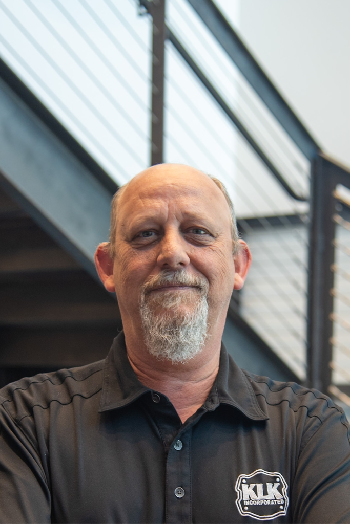 A middle-aged man with a goatee and bald head, wearing a black polo shirt, stands in front of a modern staircase with metal railings—capturing the essence of precision sheet metal manufacturing.
