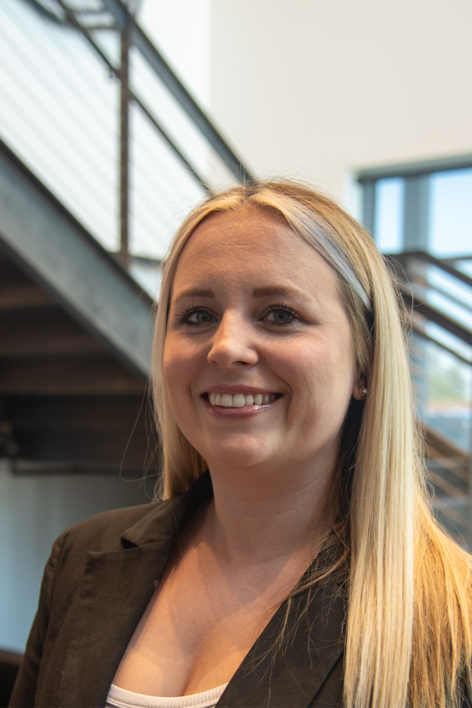 A woman with long blonde hair and a black blazer smiles in front of an industrial staircase and window, reflecting the professionalism found in Precision Sheet Metal Manufacturing.