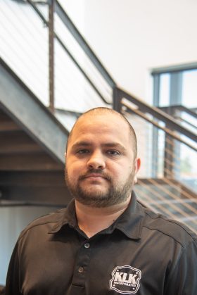 A man with short hair and beard wearing a black K1K Import Kart polo shirt stands indoors near a metal staircase.
