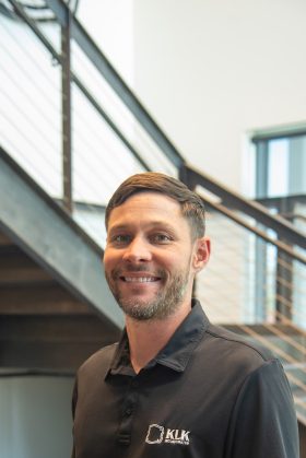 A man with short brown hair and a beard, wearing a black polo shirt with a "KLK" logo, stands indoors near a metal staircase.