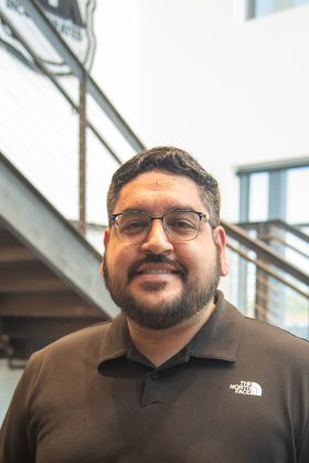 A man with glasses and a beard, wearing a black North Face polo shirt, stands indoors near a staircase and large windows.