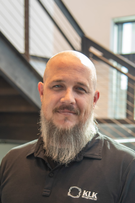 A man with a shaved head and gray beard, wearing a black polo shirt with a "KLK Incorporated" logo, stands indoors in front of a staircase and window—representing expertise in Precision Sheet Metal Manufacturing.
