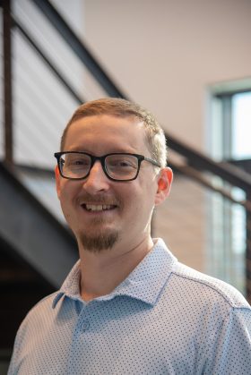 A man with short hair, glasses, and a goatee, wearing a light blue patterned shirt, stands indoors in front of a staircase, reflecting the professionalism seen in Precision Sheet Metal Manufacturing.